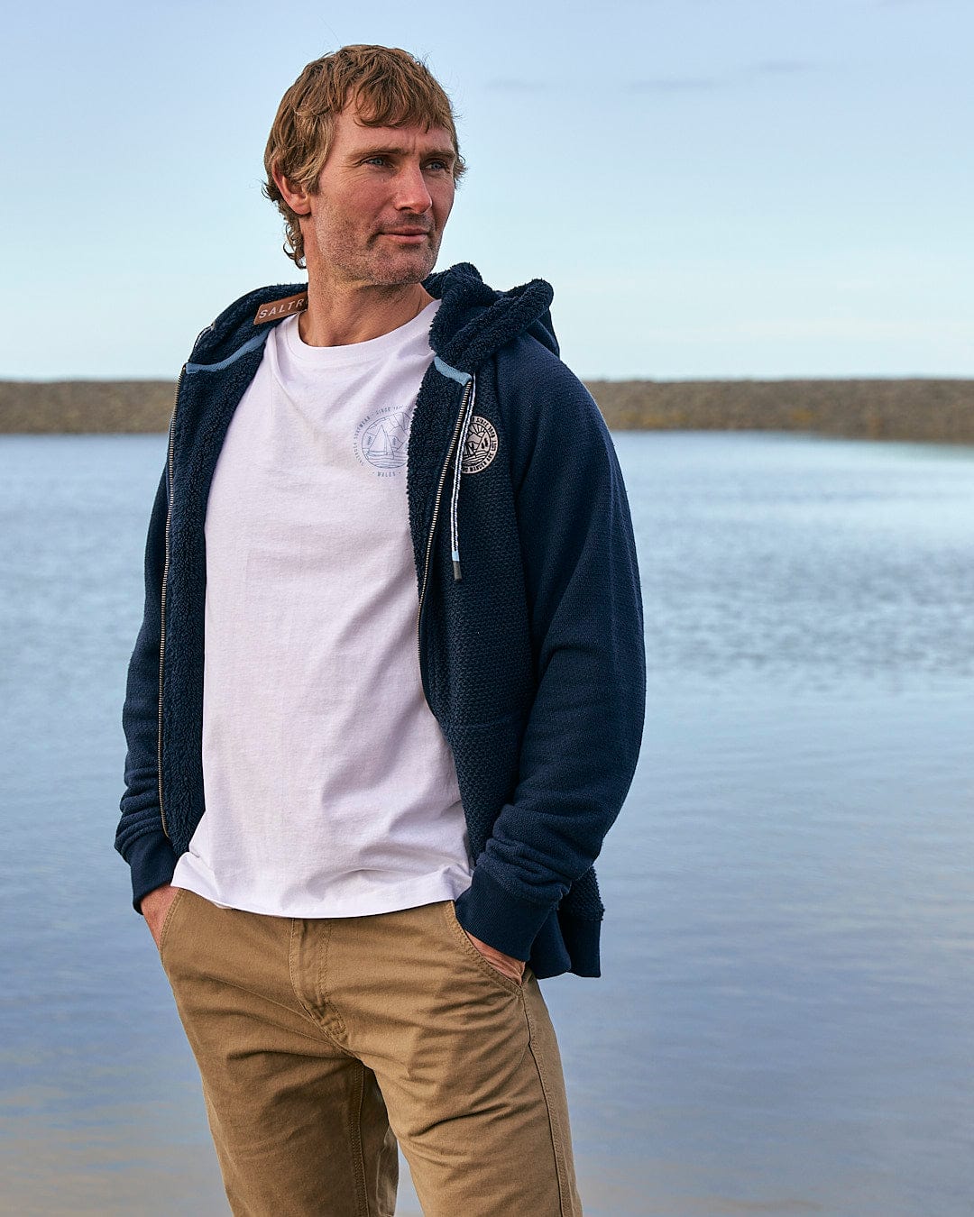 A man standing next to a body of water wearing a Saltrock Hall - Mens Orkney Borg Lined Hoodie - Dark Blue.