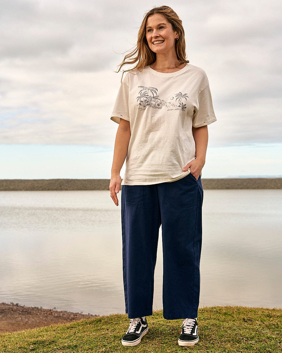 A woman is standing in front of a body of water wearing Saltrock's Hilda - Womens Canvas Trouser - Dark Blue t-shirt.