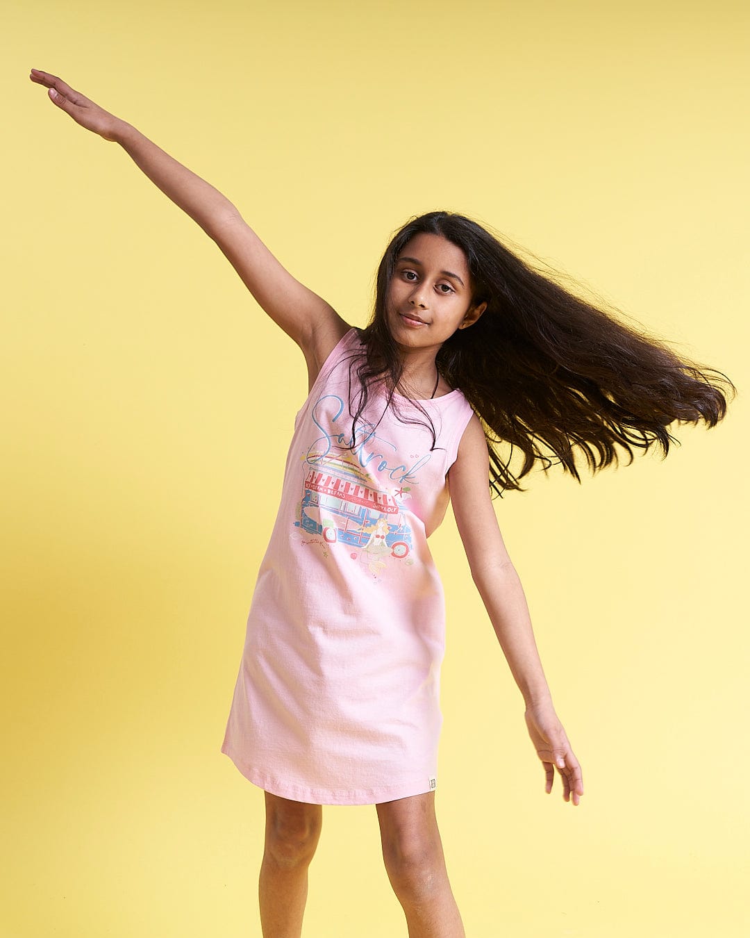 A girl in a Saltrock Ice Cream Dreams - Kids Dress - Light Pink posing on a yellow background.