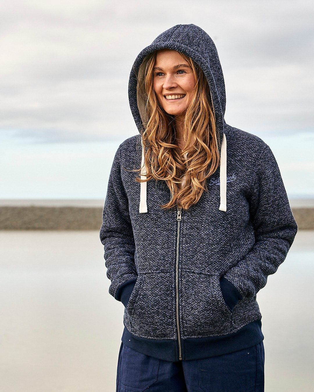 A young woman wearing a Saltrock Jazz - Womens Borg Lined Zip Hoodie - Dark Blue and jeans on the beach.