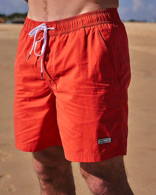 A man in Saltrock red shorts standing on the beach.