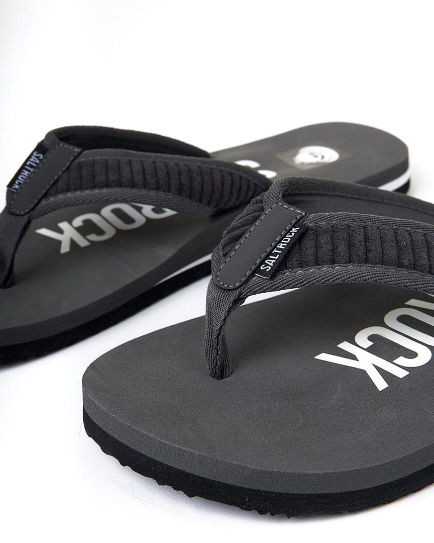 A pair of black flip flops with the word Rock Saltrock on them, replaced with the given product and brand names would be: A pair of dark grey Lineup - Cord Flip Flops by Saltrock.