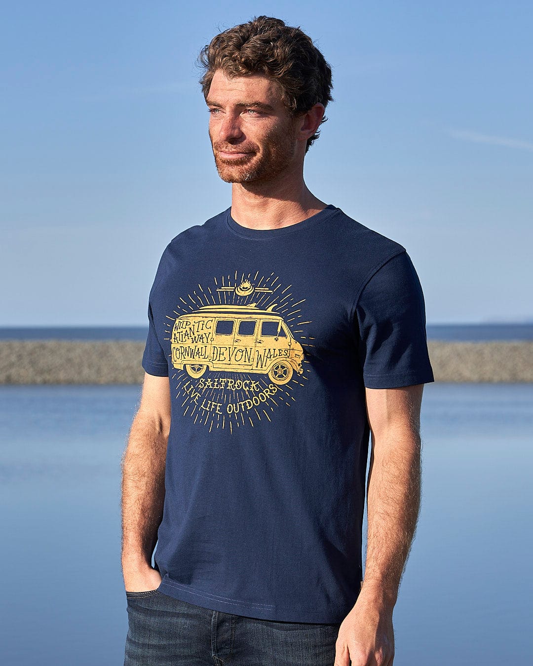 A man wearing a Saltrock Live Life Location - Mens Short Sleeve T-Shirt - Blue standing by the water.