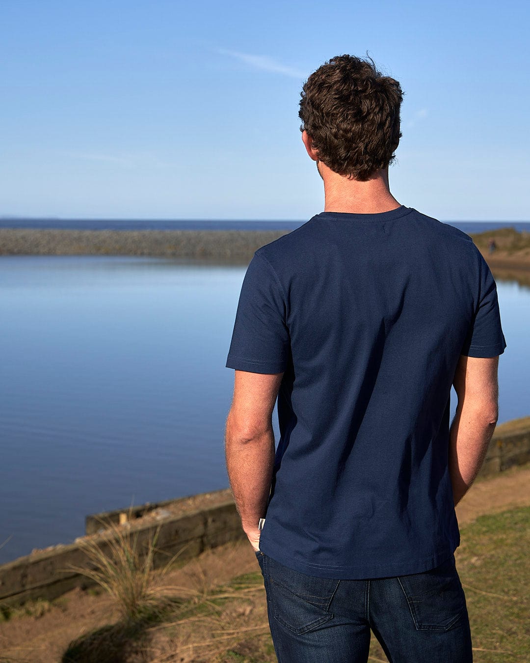 The Saltrock Live Life Location - Mens Short Sleeve T-Shirt - Blue is being worn by a man looking at a body of water.