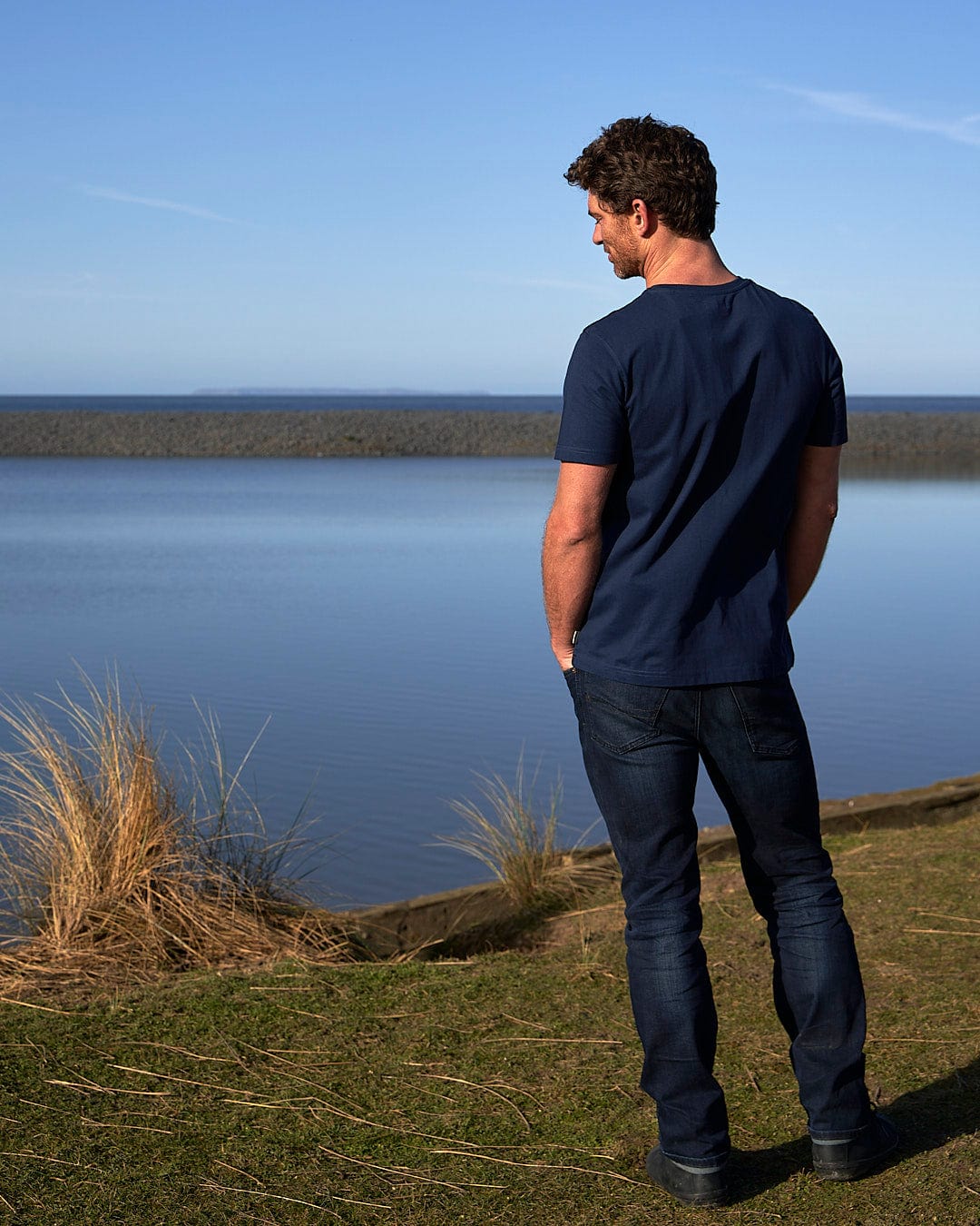 A man is standing in front of a body of water, wearing the Saltrock Live Life Location - Mens Short Sleeve T-Shirt - Blue.