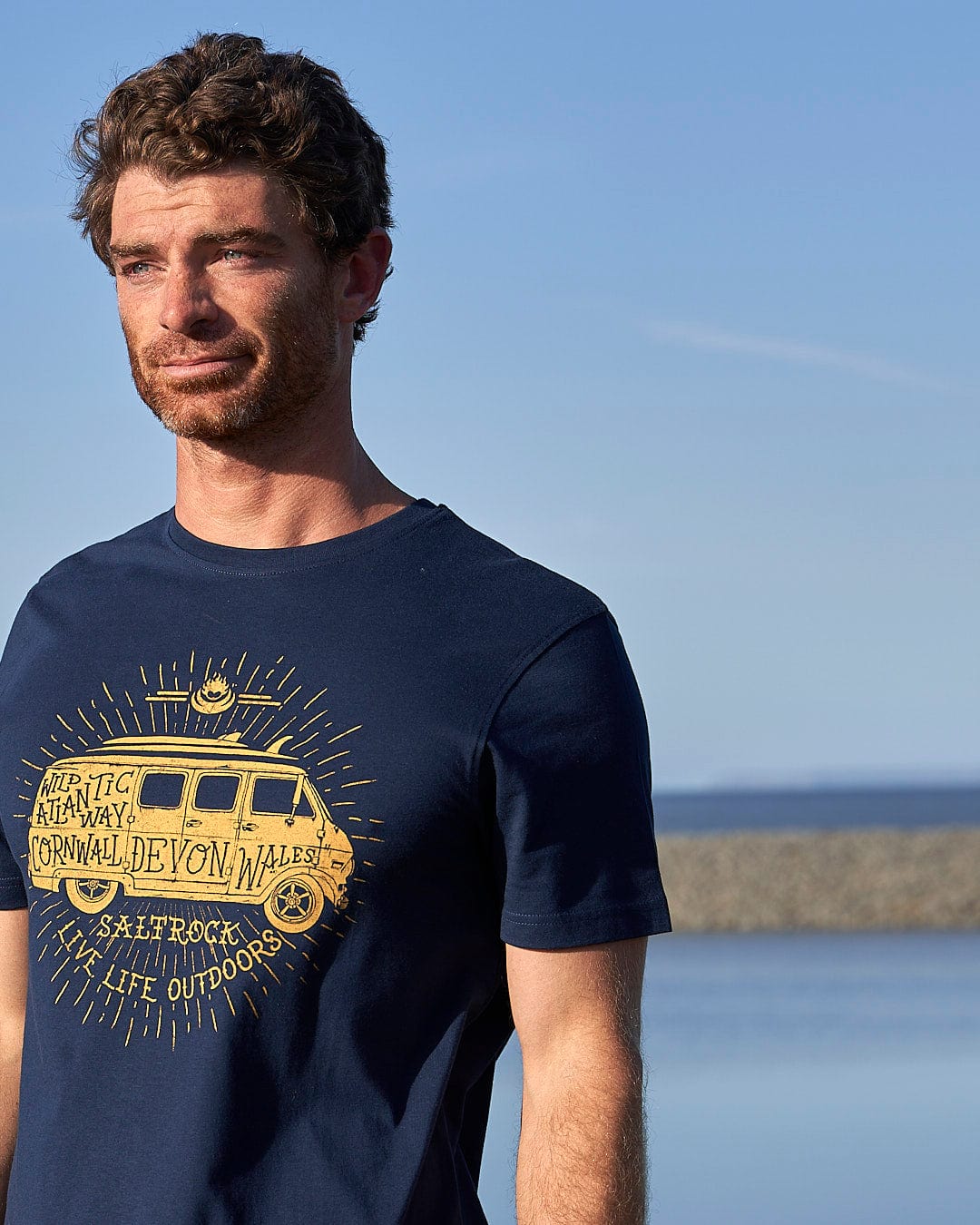A man wearing a Saltrock Live Life Location - Mens Short Sleeve T-Shirt - Blue with a van on it.