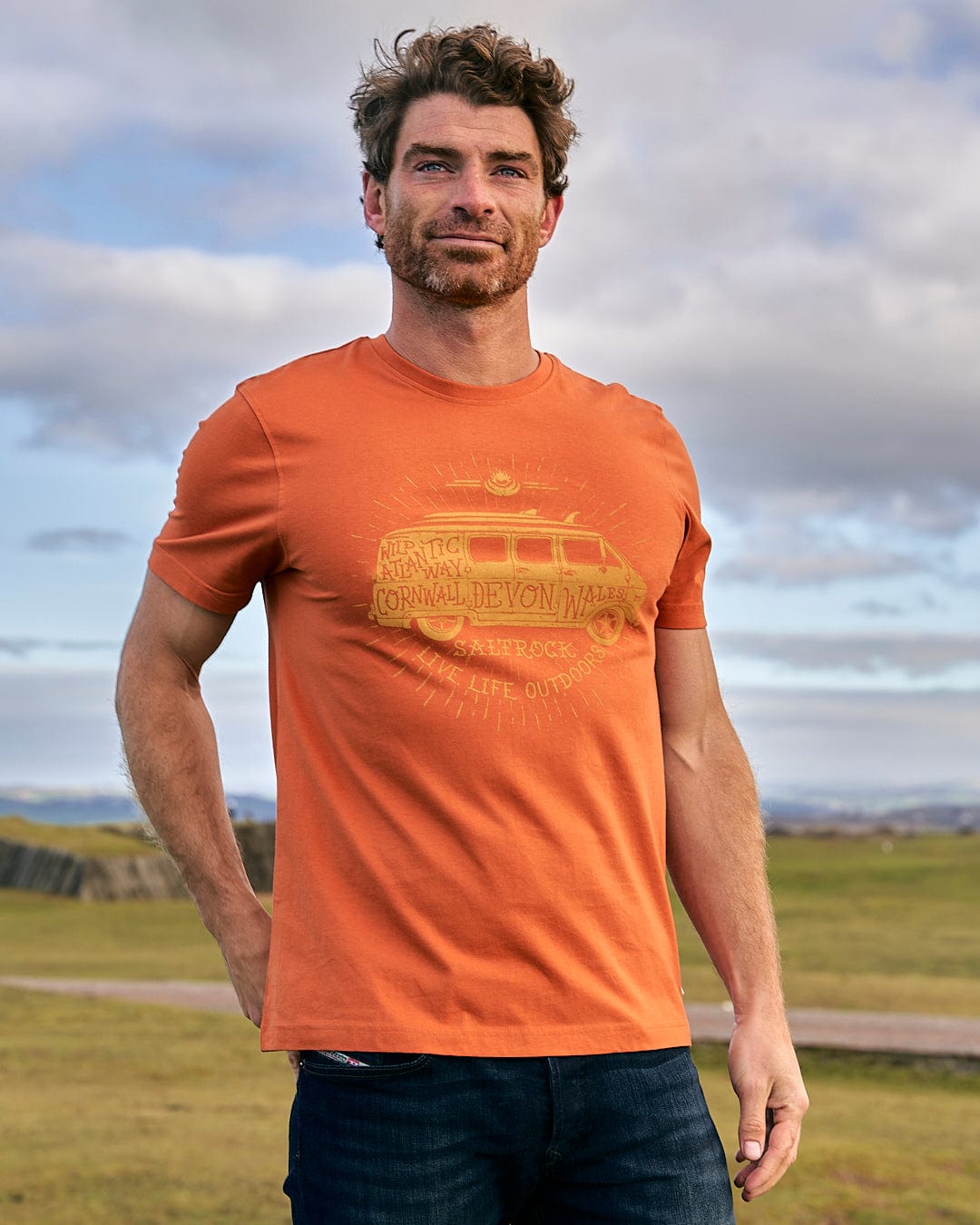 A man standing in a field wearing a Saltrock Live Life Location - Mens Short Sleeve T-Shirt - Orange.