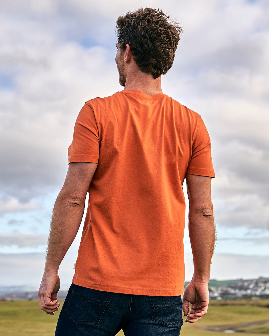 The back of a man in a Saltrock Live Life Location - Mens Short Sleeve T-Shirt - Orange standing in a field.