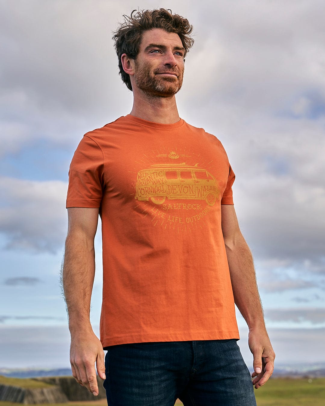 A man wearing a Saltrock Live Life Location - Mens Short Sleeve T-Shirt - Orange standing in a field.