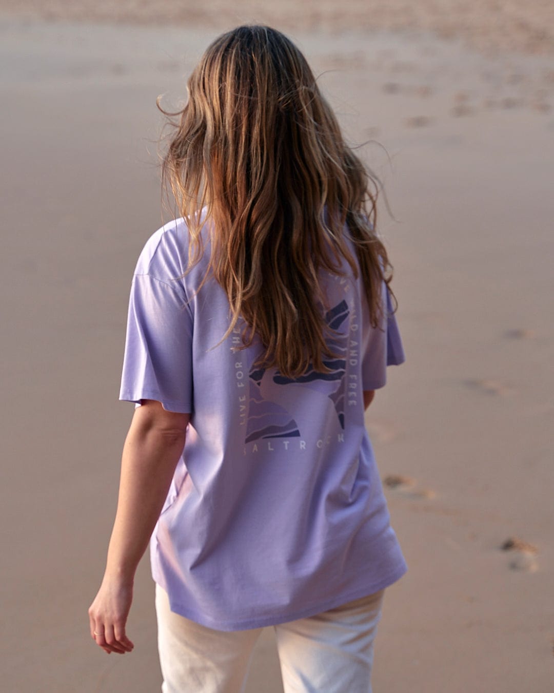 A woman walking on the beach wearing a Saltrock Live Wild - Womens Short Sleeve T-Shirt - Light Purple.