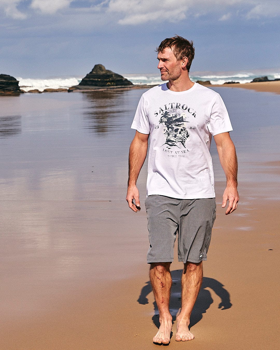 A man walking on a beach wearing the Lost At Sea Skull - Mens Short Sleeve T-Shirt in White by Saltrock.