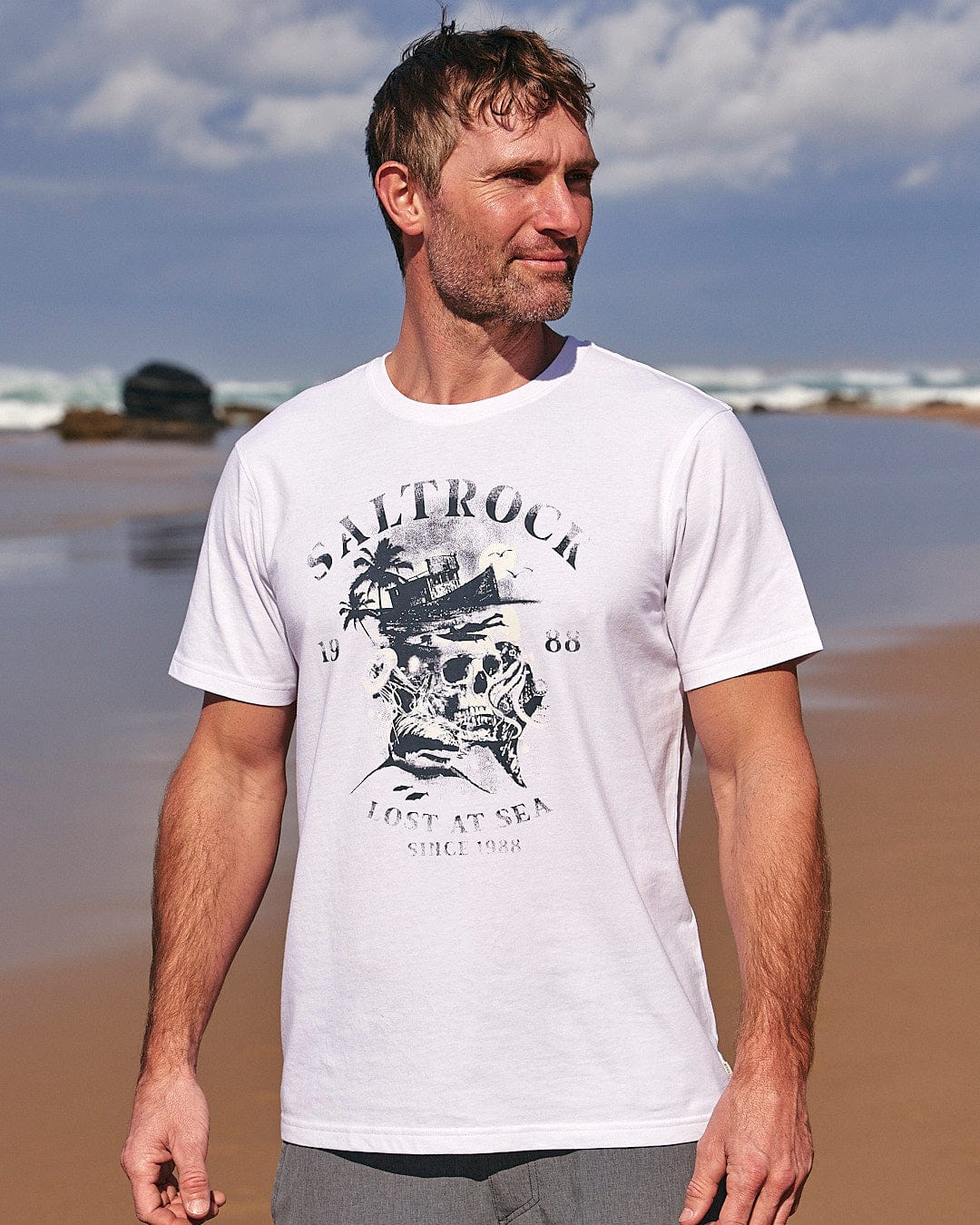 A man standing on the beach wearing a Lost At Sea Skull - Mens Short Sleeve T-Shirt - White by Saltrock.