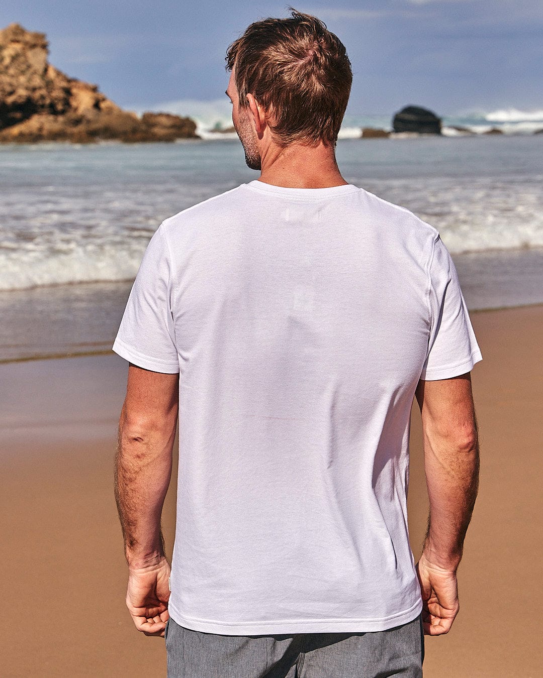 A man standing on the beach wearing a Saltrock Lost At Sea Skull - Mens Short Sleeve T-Shirt - White.