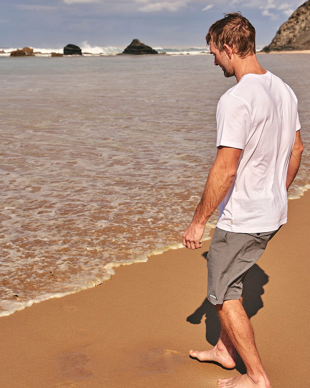 A man standing on a beach with a Saltrock Lost At Sea Skull - Mens Short Sleeve T-Shirt - White surfboard.