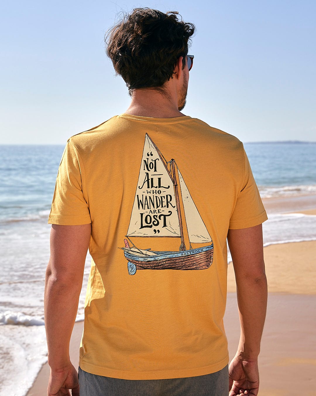A man standing on the beach wearing a Lost Ships - Mens Short Sleeve T-Shirt in Yellow by Saltrock with a sailboat on it.