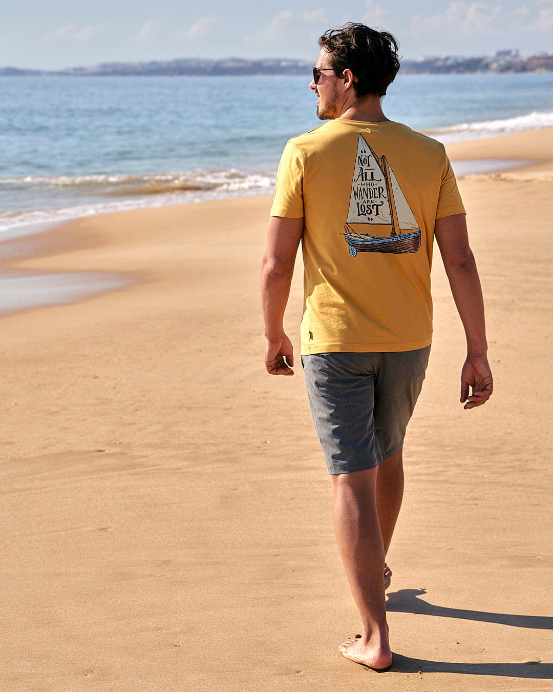 A man walking on a beach with a Saltrock Lost Ships - Mens Short Sleeve T-Shirt - Yellow on his shirt.