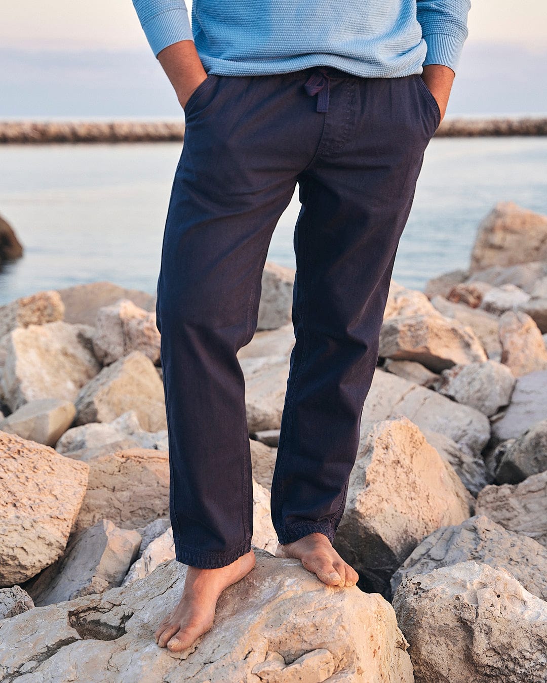 A man enjoying an outdoor adventure, standing on rocks next to the ocean, confidently donning a Meddon - Mens Twill Trouser - Navy by Saltrock.