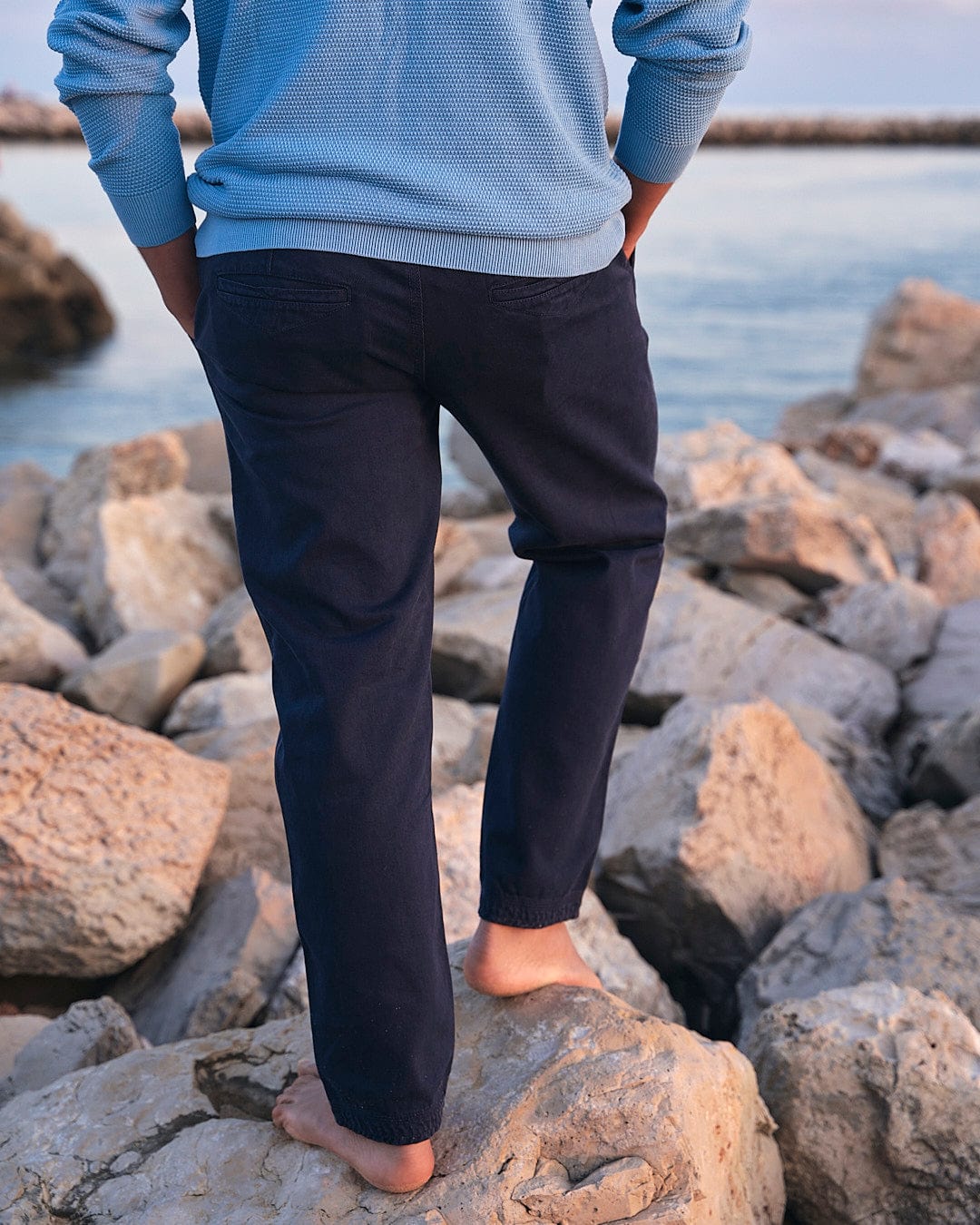A man wearing Saltrock Meddon - Mens Twill Trouser - Navy cuffed trousers standing on rocks near the ocean.