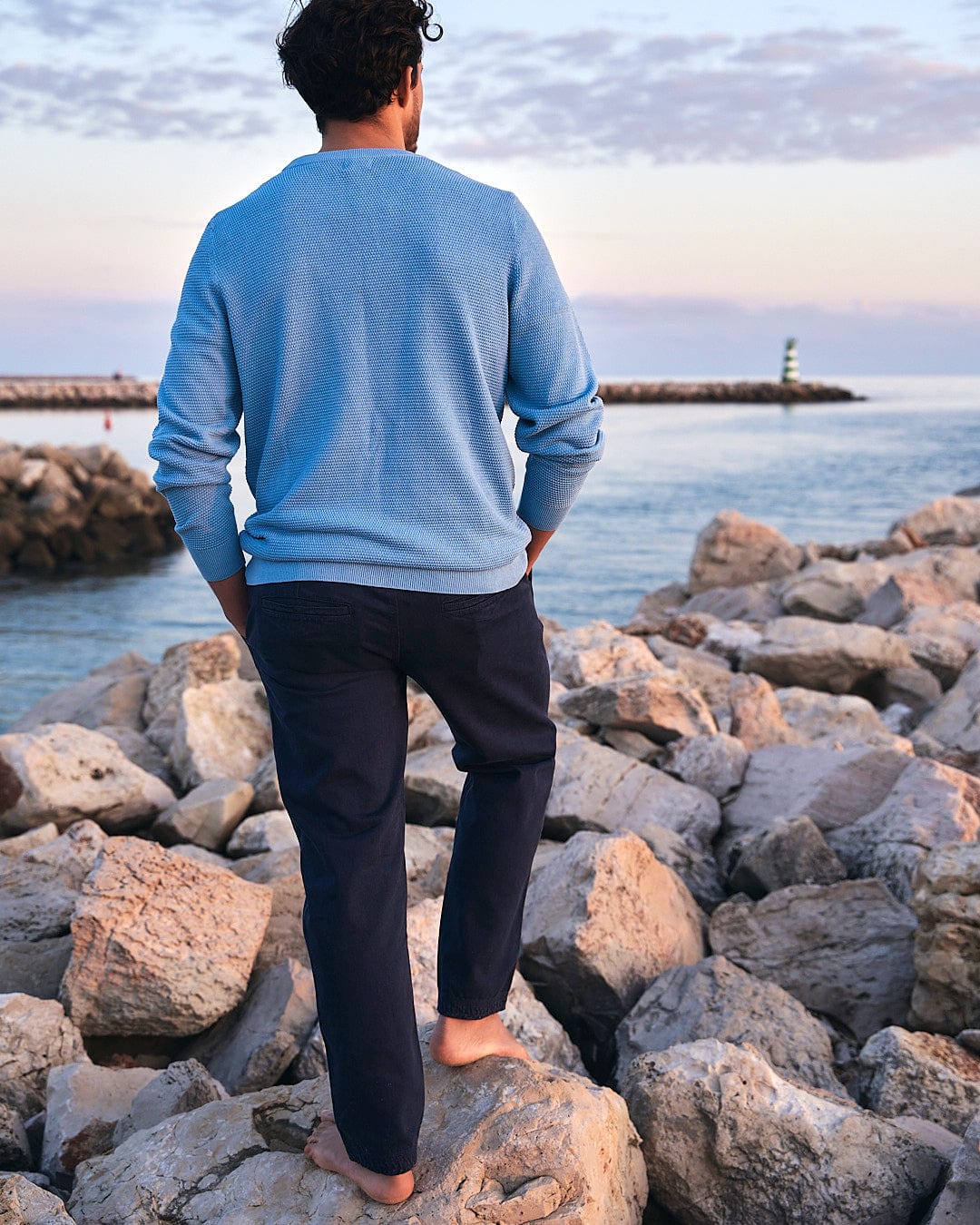 A man wearing Saltrock Meddon - Mens Twill Trouser - Navy cuffed twill trousers, standing on rocks, mesmerized by the vastness of the ocean during his outdoor adventure.