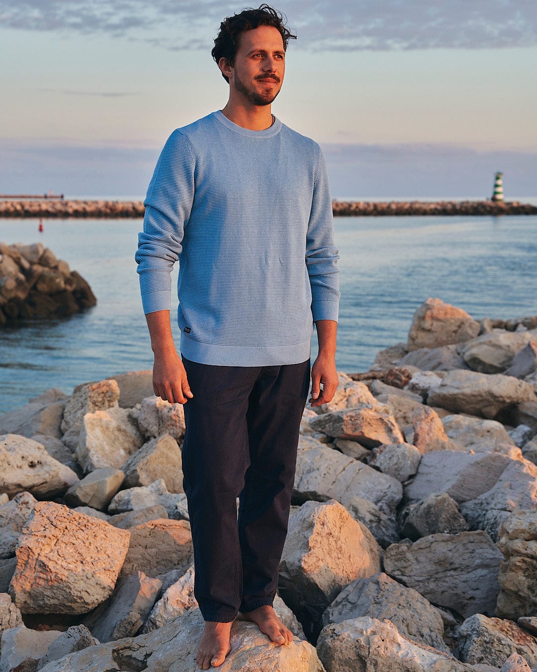 An outdoor adventurer standing on rocks next to the ocean wearing a blue sweater and Saltrock Meddon - Mens Twill Trouser - Navy.