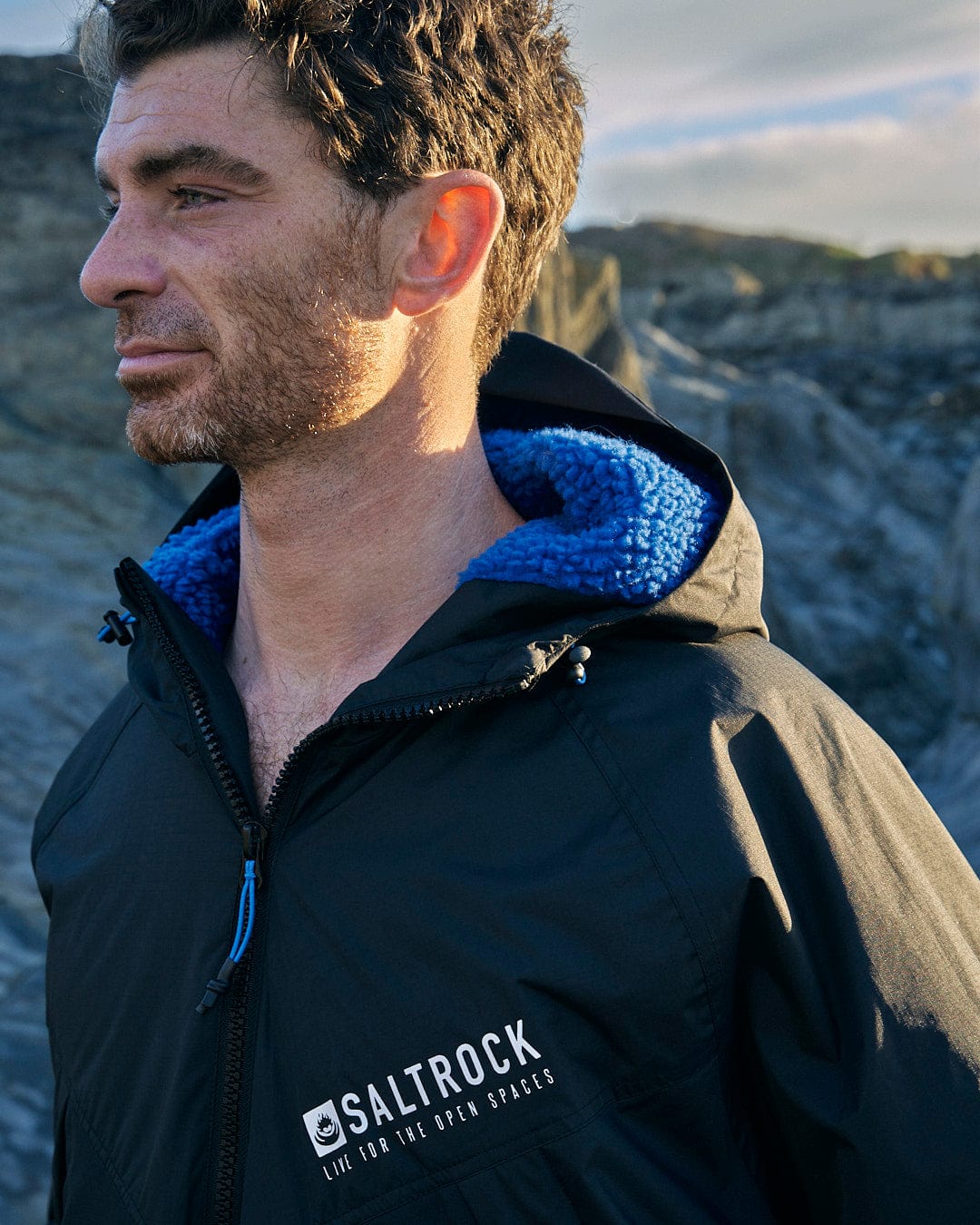 A man is standing in front of a rocky area wearing a Saltrock Four Seasons - Waterproof Changing Robe in Black/Blue.