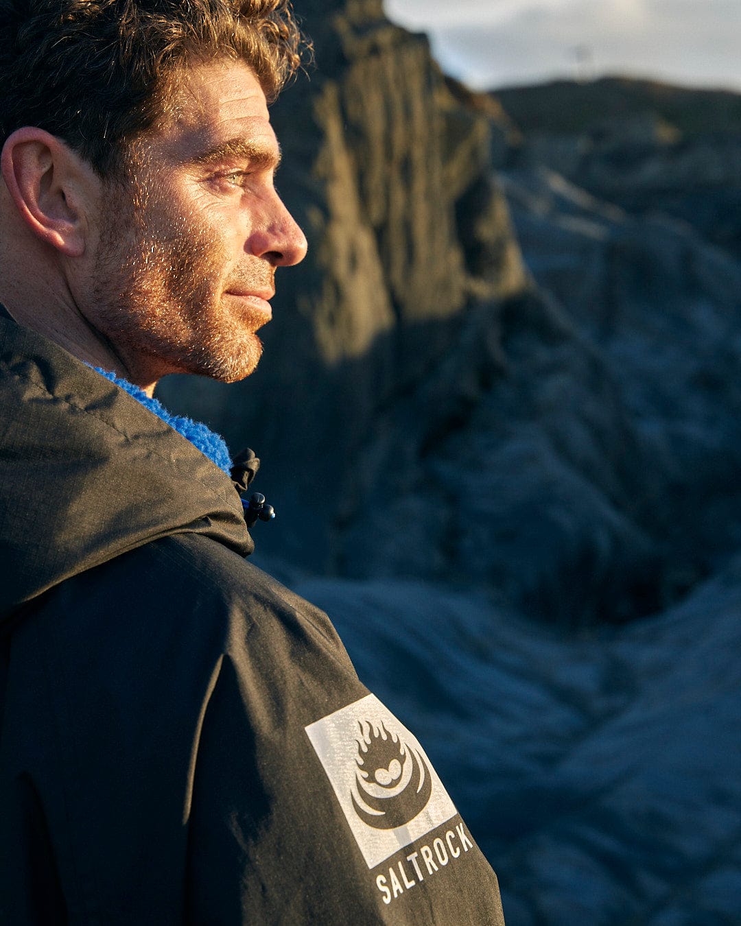 A man in a Saltrock black jacket standing in front of a cliff.