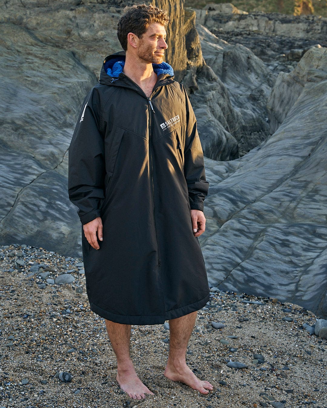 A man in a Saltrock black hooded coat, the Four Seasons Waterproof Changing Robe in Black/Blue, standing on a rocky beach.