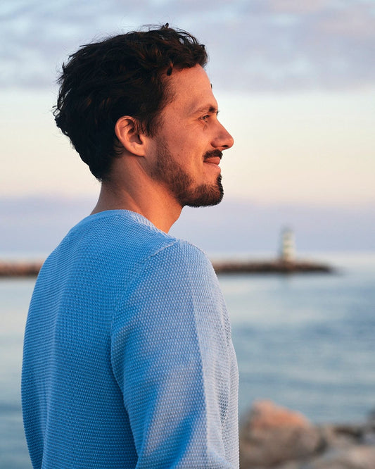 A man with a beard looking at the ocean wearing Moss - Mens Washed Knitted Crew - Light Blue by Saltrock.