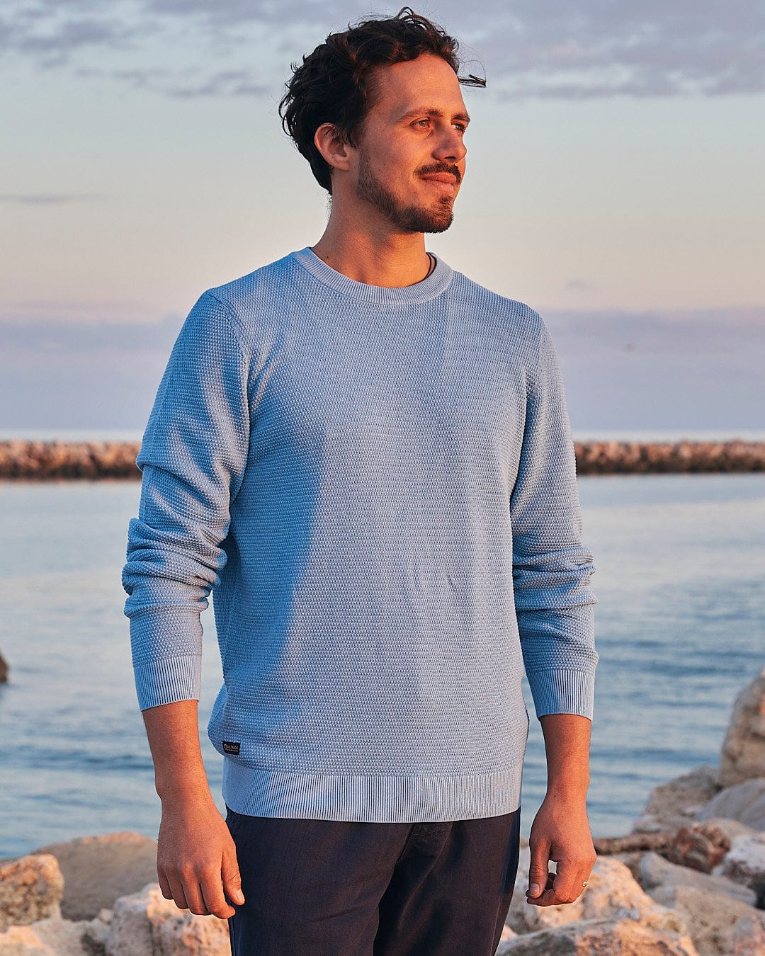 A man in a Saltrock - Moss - Mens Washed Knitted Crew - Light Blue sweater standing by the ocean.