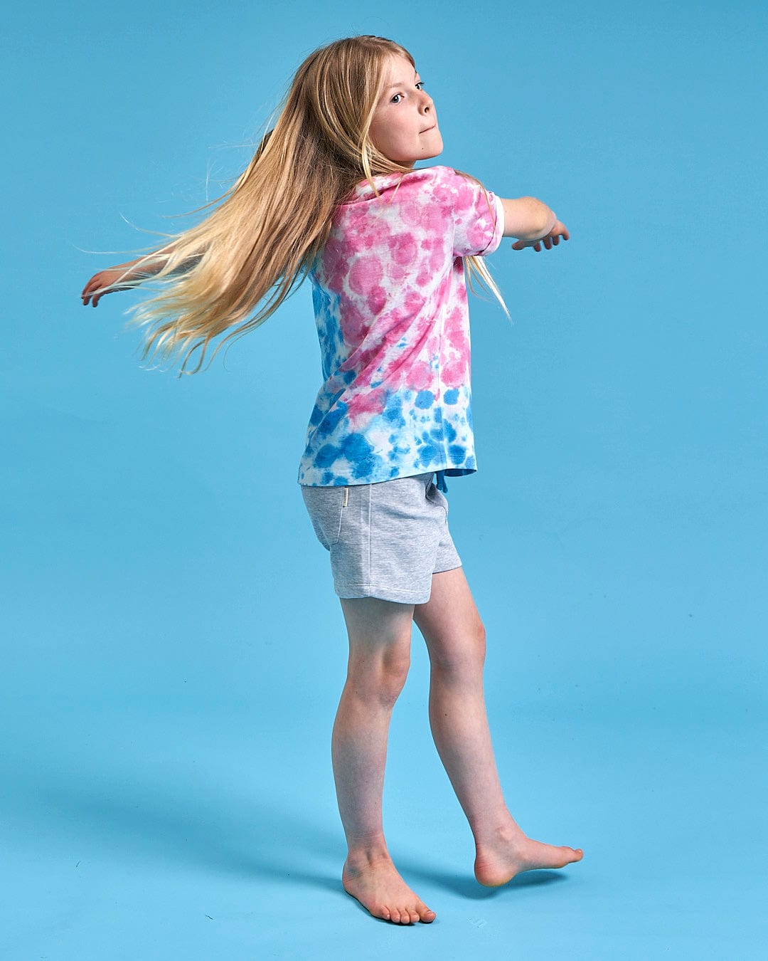 A girl wearing a tie dye Saltrock Mermaid Surf - Kids Tie Dye Short Sleeve T-Shirt - Blue/Pink is standing on a blue background, beach ready.