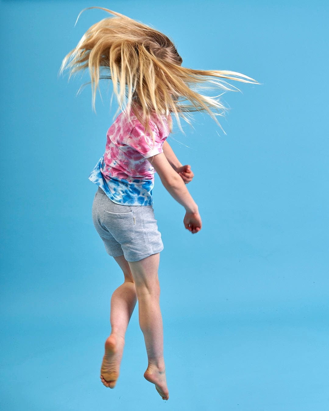 A beach ready young girl jumping in the air with her hair blowing in the wind, wearing a Saltrock Mermaid Surf - Kids Tie Dye Short Sleeve T-Shirt - Blue/Pink.