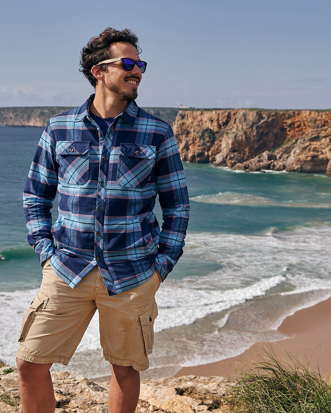 A man in a Saltrock Ocean - Mens Borg Lined Shirt - Blue Check is standing on a rocky beach, gazing at the ocean.