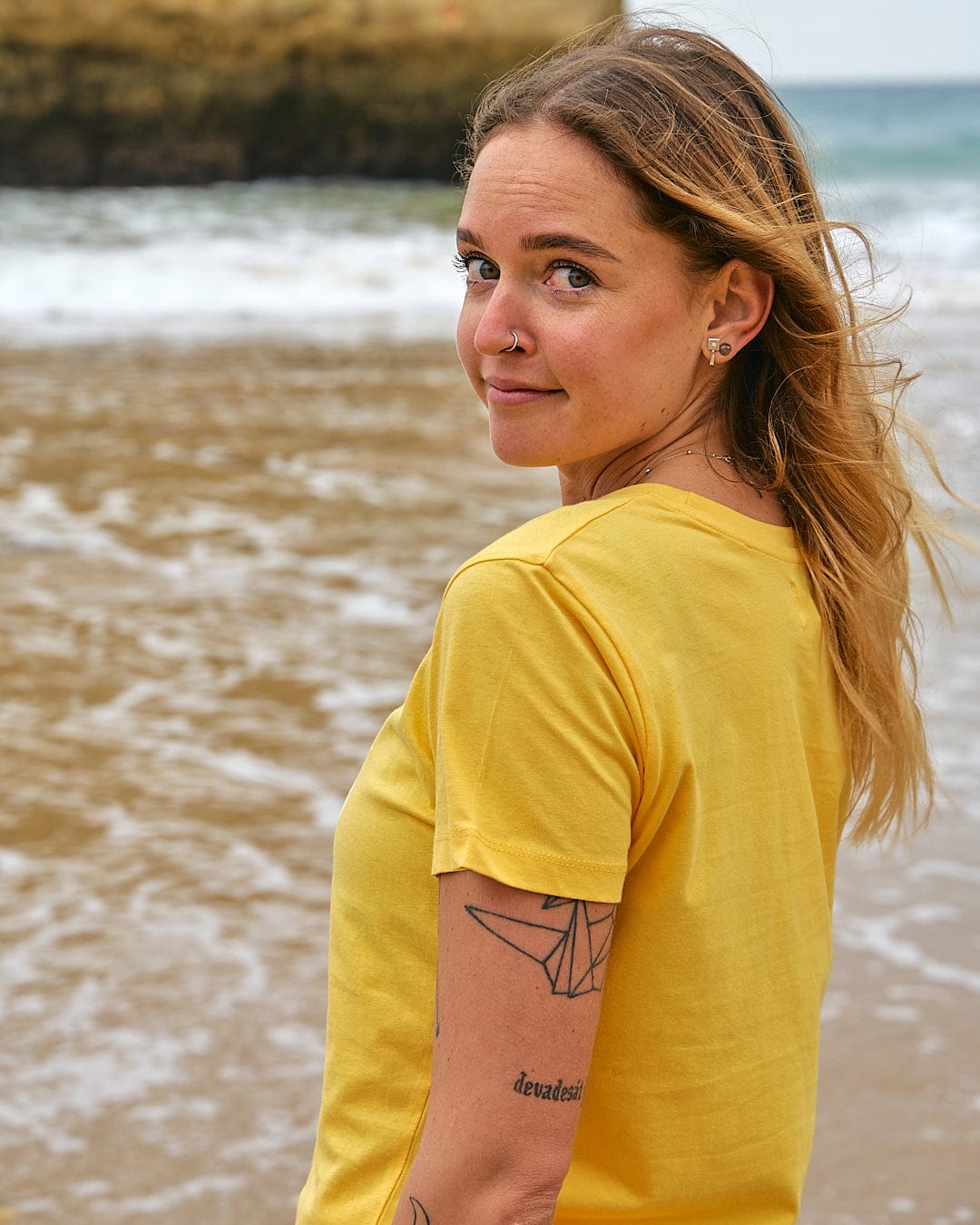 A woman wearing the Saltrock On The Road - Womens Short Sleeve T-Shirt - Yellow standing on the beach.