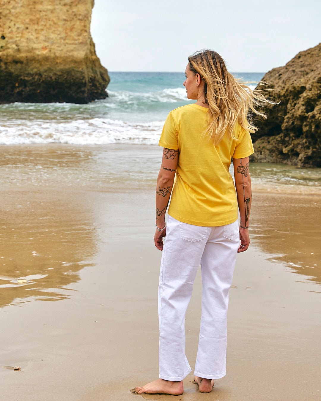 A woman wearing the Saltrock On The Road - Womens Short Sleeve T-Shirt - Yellow standing on a beach.