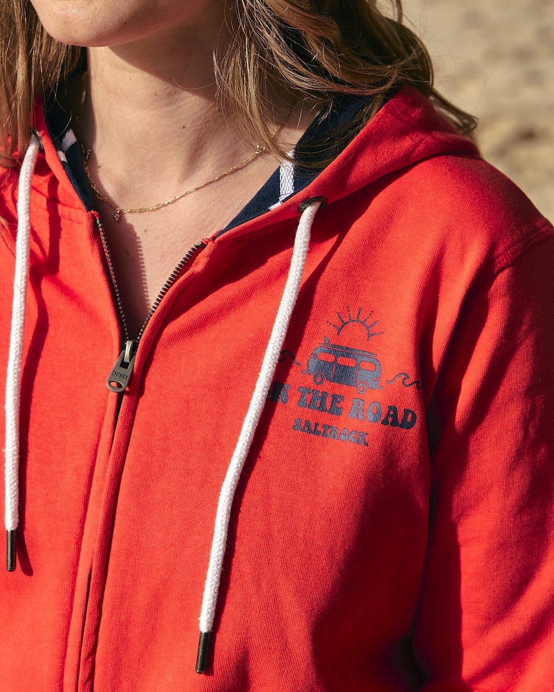 A woman wearing a Saltrock red On The Road - Womens Zip Hoodie on the beach.