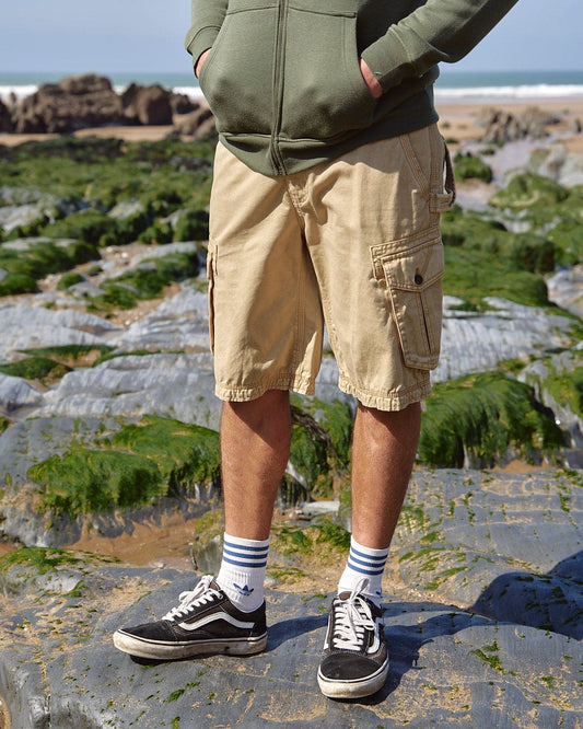 A person standing on a rocky shore wearing Saltrock's Penwith II - Mens Cargo Shorts in Cream with cargo pockets, sneakers, and socks.