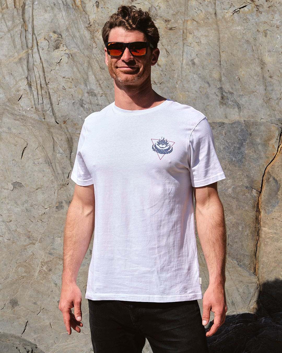 A man wearing sunglasses and a Saltrock Poolside Wave - Mens Short Sleeve T-Shirt - White standing in front of a rock.