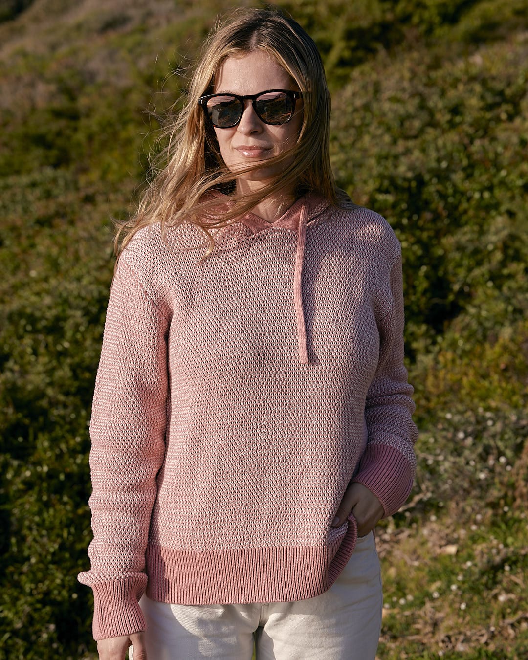 A woman in a Saltrock Poppy - Womens Knitted Pop Hoodie - Mid Pink standing in a field.