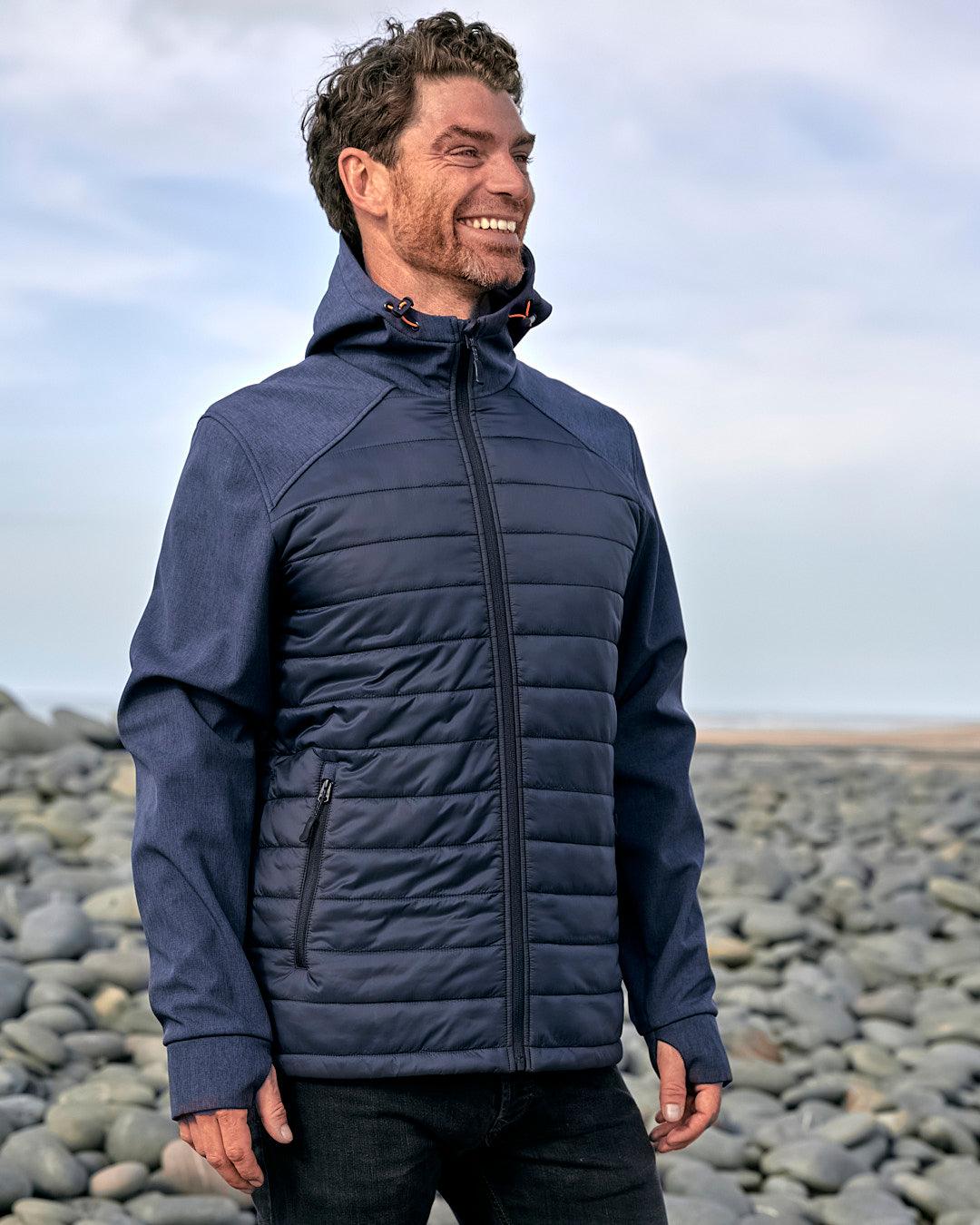 A man in a Saltrock Purbeck Padded Jacket - Dark Blue standing on a rocky beach.