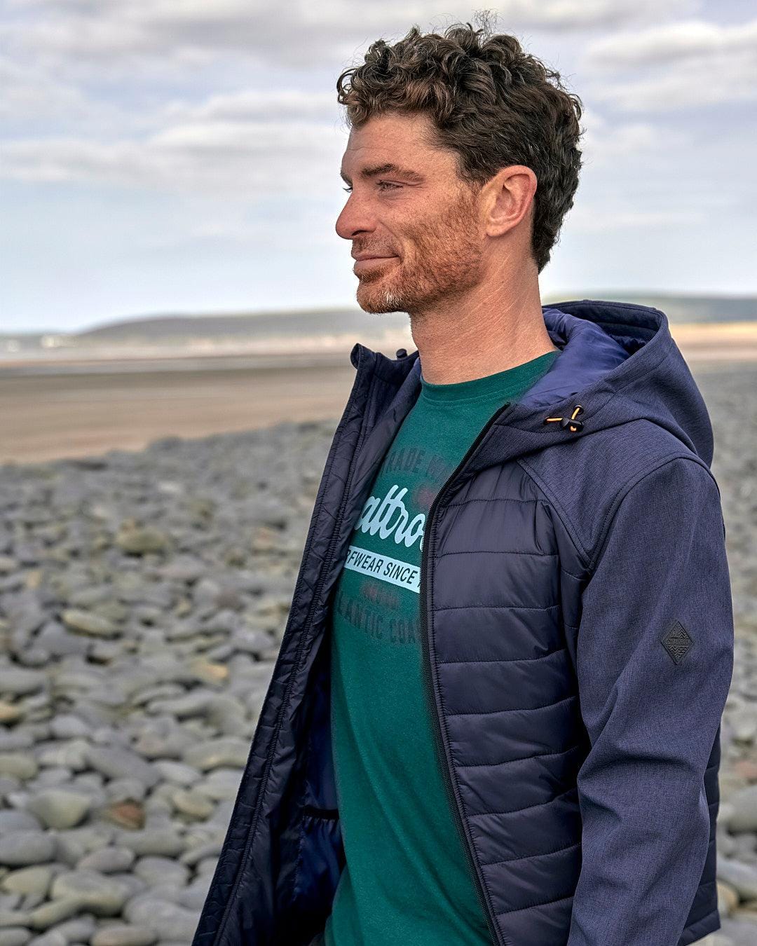 A man standing on a water-resistant beach wearing a Saltrock Purbeck Padded Jacket - Dark Blue.
