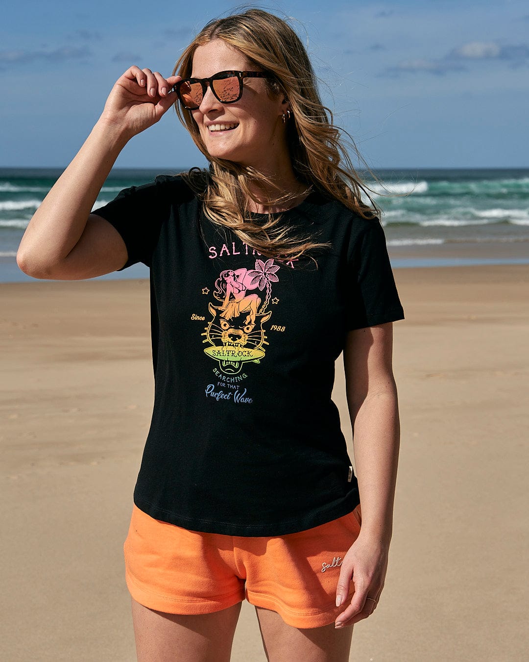 A woman wearing a Saltrock Purfect Wave Gradient - Womens Short Sleeve T-Shirt - Black and orange shorts standing on the beach.