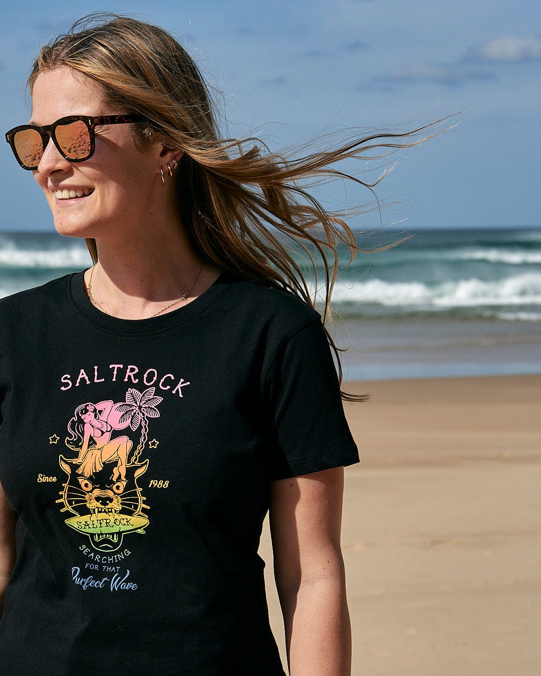 A woman is standing on the beach wearing a Saltrock t-shirt that says Purfect Wave Gradient - Womens Short Sleeve T-Shirt - Black.