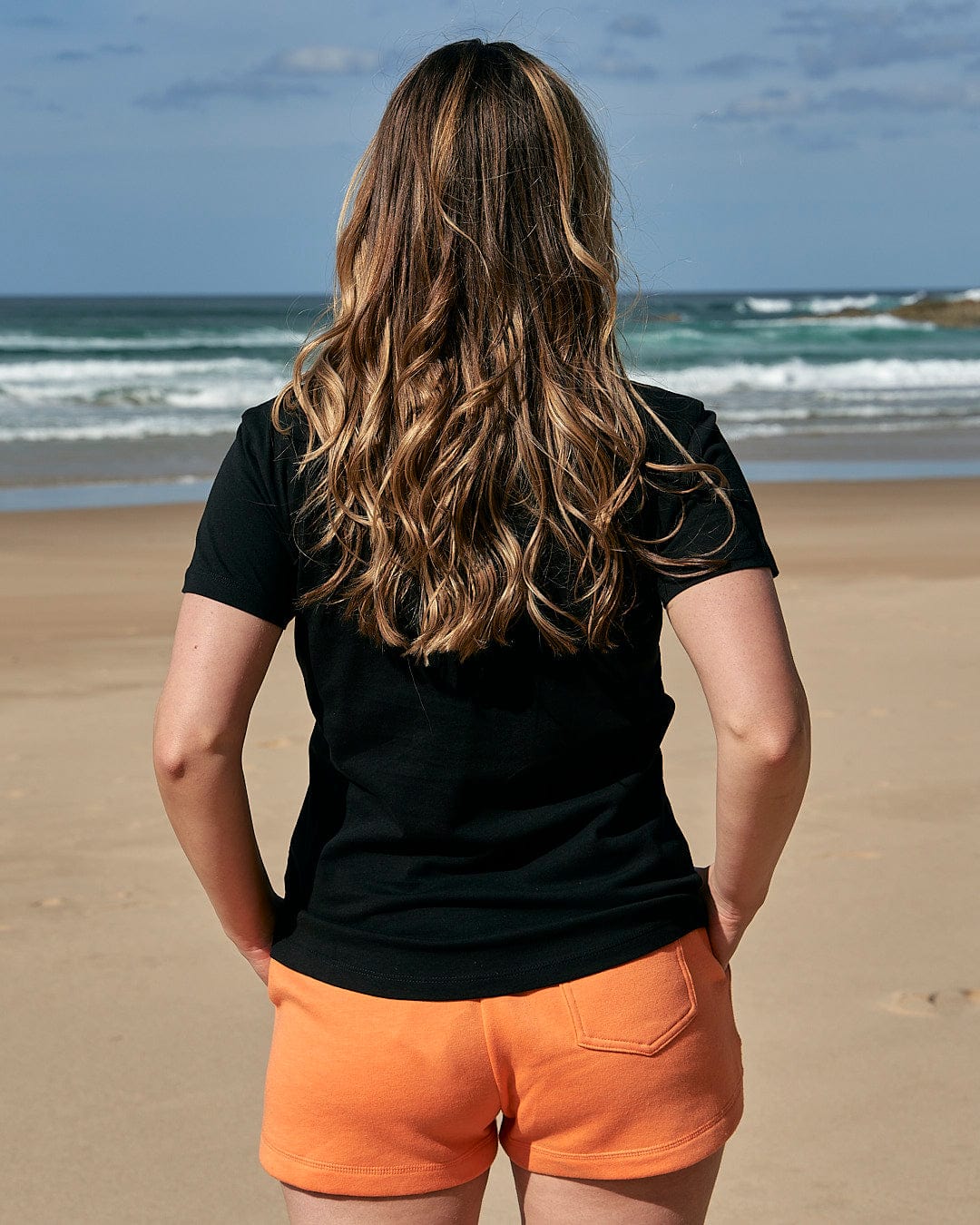 A woman wearing a Saltrock Purfect Wave Gradient - Womens Short Sleeve T-Shirt - Black and orange shorts on the beach.