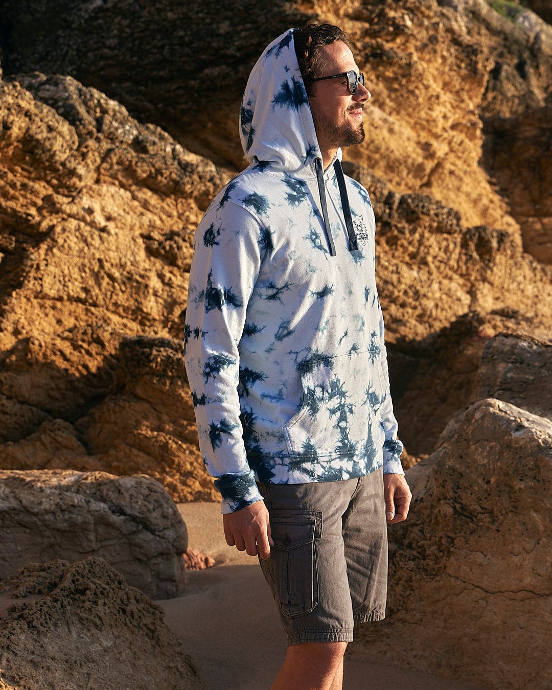 A man wearing a Saltrock Purfect Wave - Mens Tie Dye Pop Hoodie - Light Blue standing on a rocky beach.