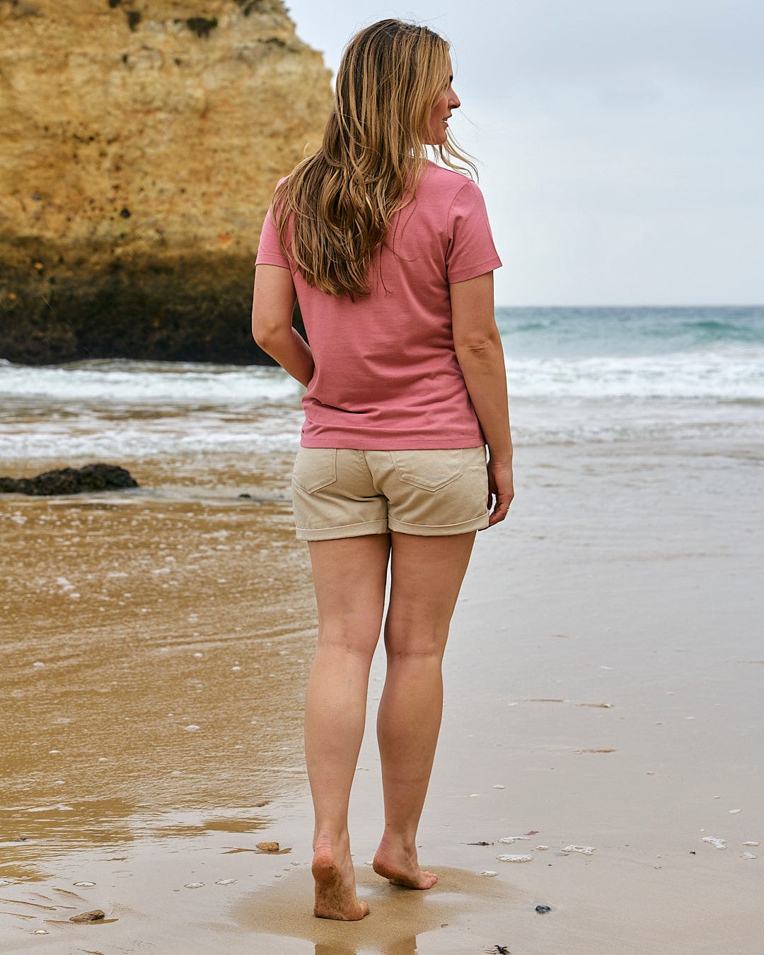 A woman standing on a beach wearing a Saltrock Love Your Ocean - Womens Short Sleeve T-Shirt - Mid Pink shirt and tan shorts.