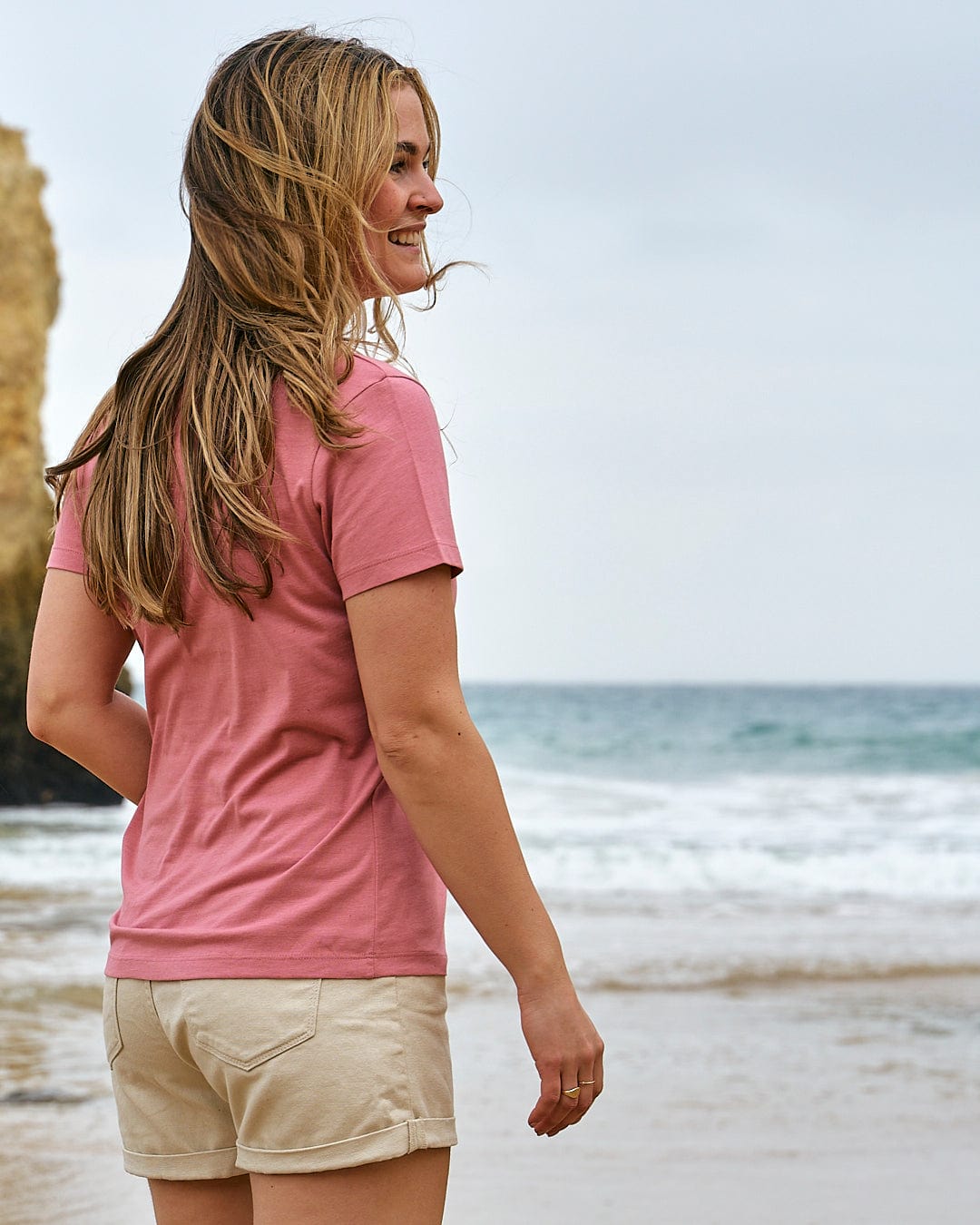 A woman in a pink shirt standing on a beach wearing the Love Your Ocean - Womens Short Sleeve T-Shirt - Mid Pink by Saltrock.