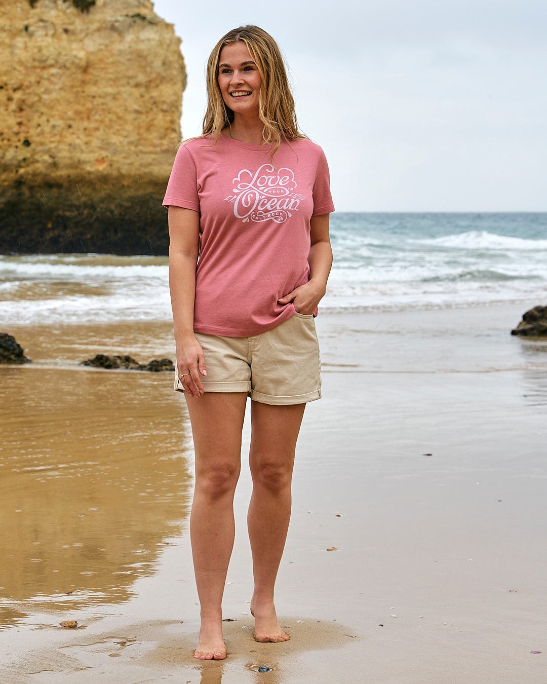 A woman standing on the beach wearing a Love Your Ocean - Womens Short Sleeve T-Shirt - Mid Pink by Saltrock.