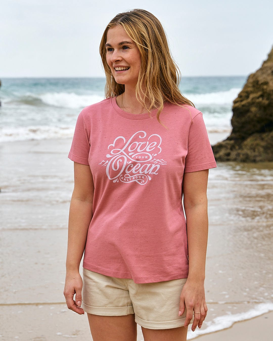 A woman is standing on the beach wearing a Saltrock Love Your Ocean - Womens Short Sleeve T-Shirt - Mid Pink.