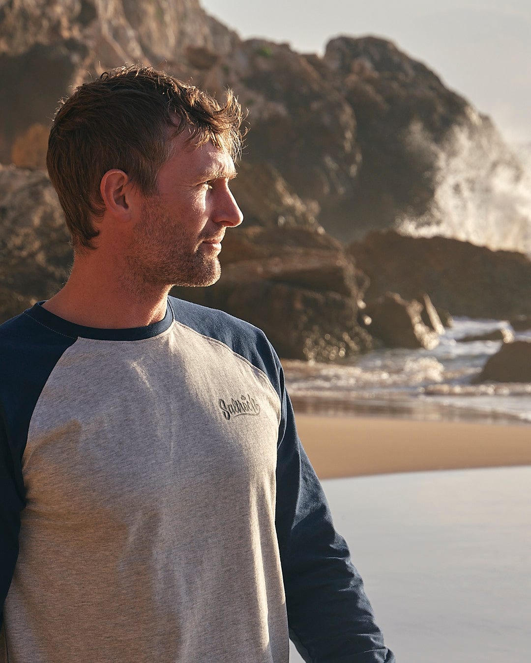 A man standing on a beach with a Saltrock Speed - Mens Long Sleeve Raglan T-Shirt - Blue surfboard.