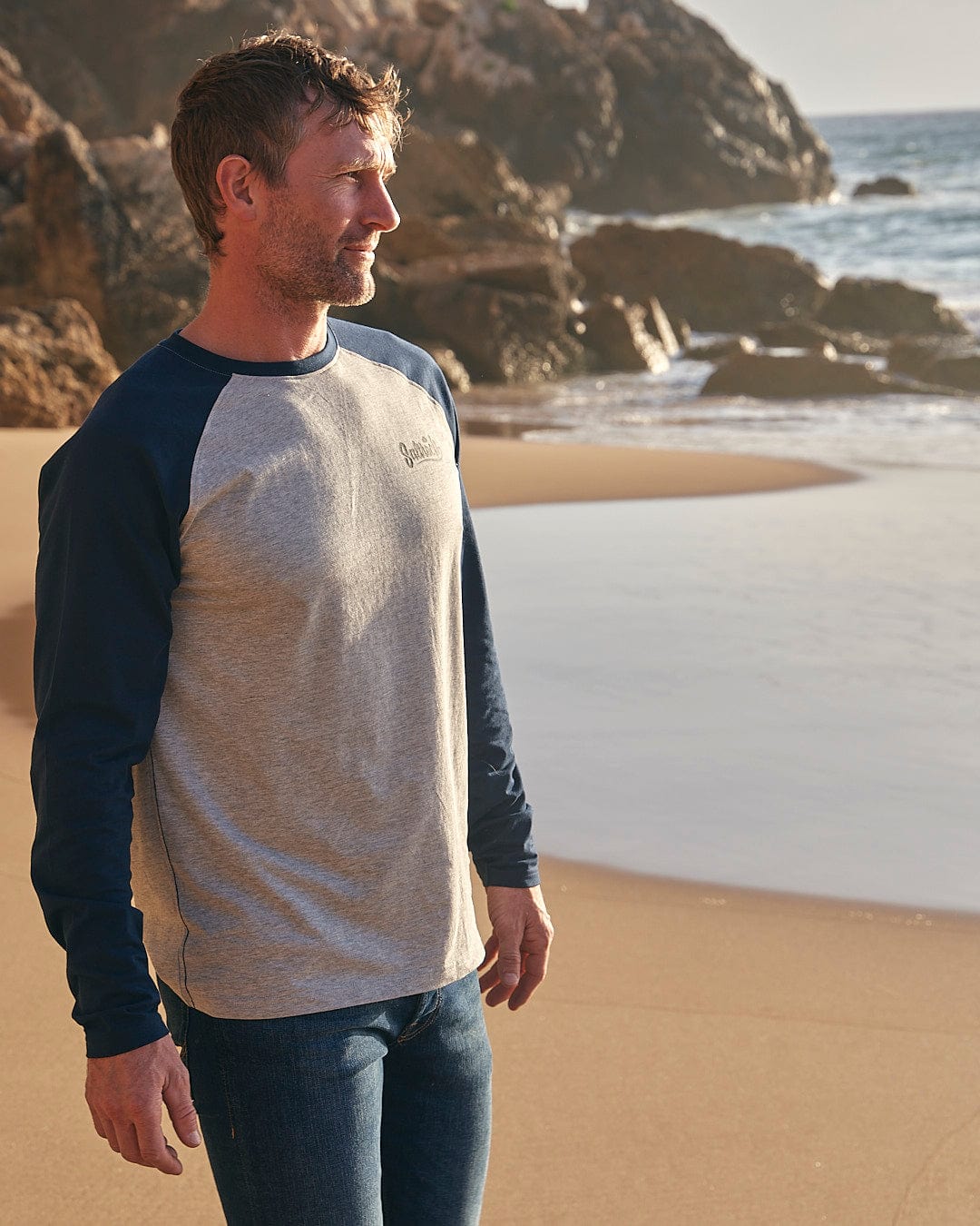 A man standing on a beach wearing Speed - Mens Long Sleeve Raglan T-Shirt - Blue by Saltrock jeans and a t-shirt.